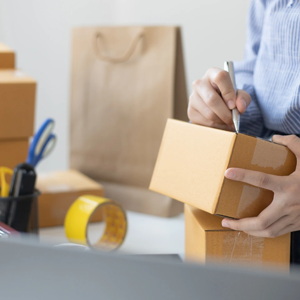 person-in-blue-shirt-writing-on-small-cardboard-box-with-packaging-supplies-and-brown-bag-nearby
