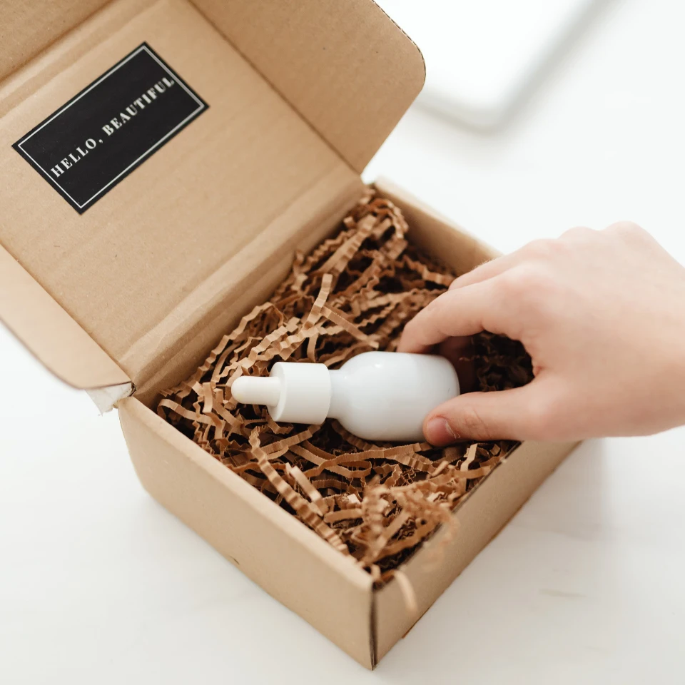 hand-removing-white-cosmetic-products-from-cardboard-box-with-brown-paper-filling-and-hello-beautiful-label