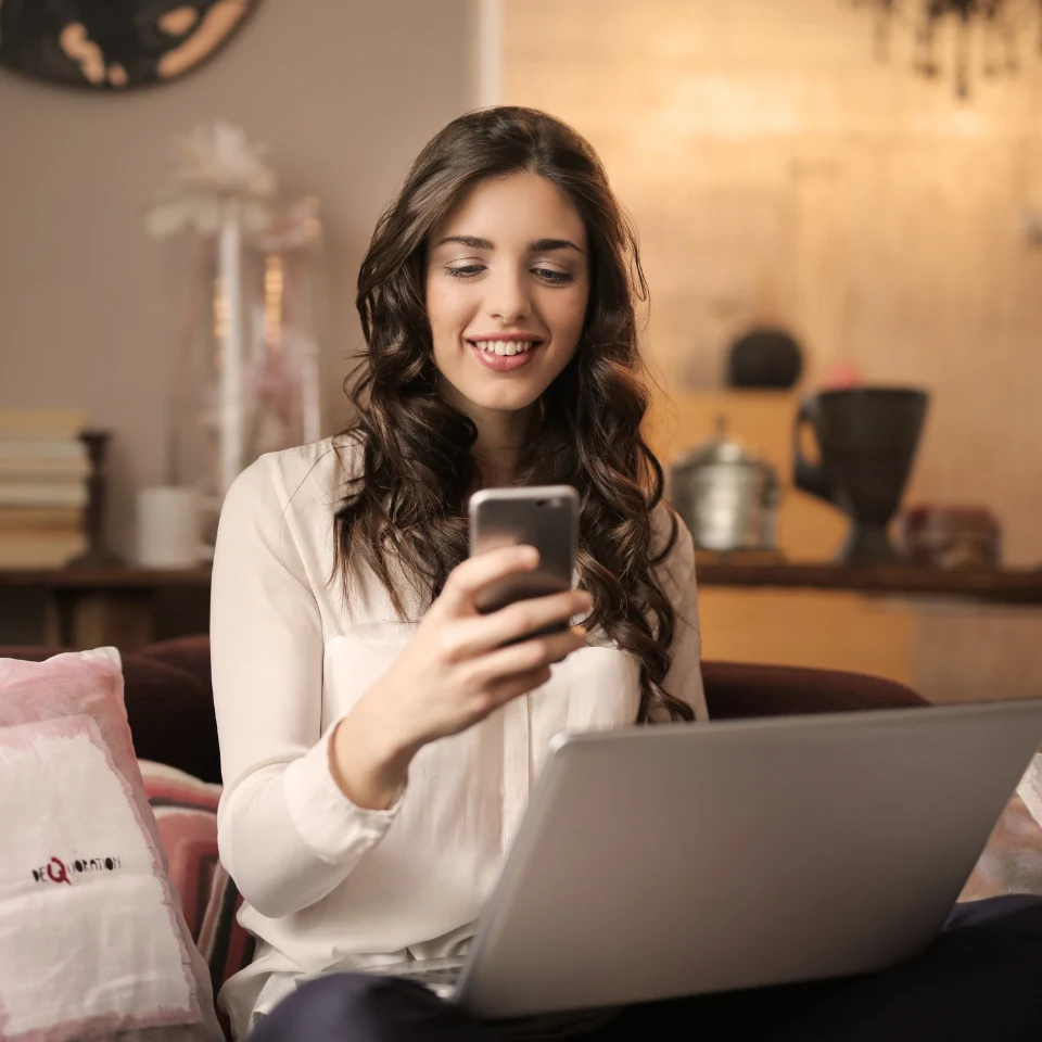 smiling-woman-in-white-sweater-using-smartphone-and-laptop-sitting-on-couch-at-home