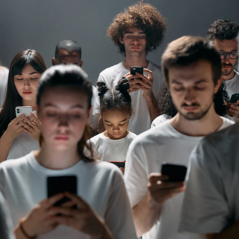 Group of diverse people wearing white shirts, standing closely together and looking down at their smartphones, with serious or neutral expressions.