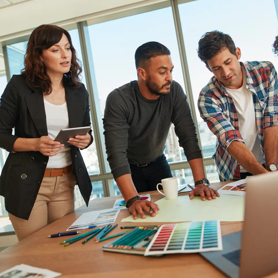 Creative team collaborating around a table with color swatches, drawings, and a laptop in a modern office during a design meeting