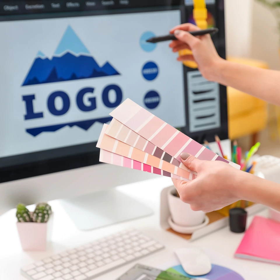 hands-holding-pink-color-palette-swatches-while-designing-blue-mountain-logo-on-computer-screen