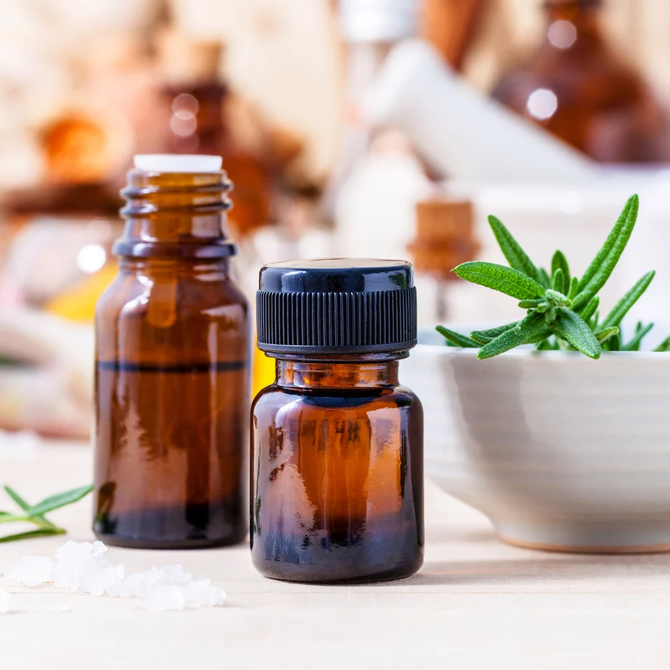 Two amber glass essential oil bottles next to a white bowl filled with fresh rosemary and coarse salt, on a natural wood table.
