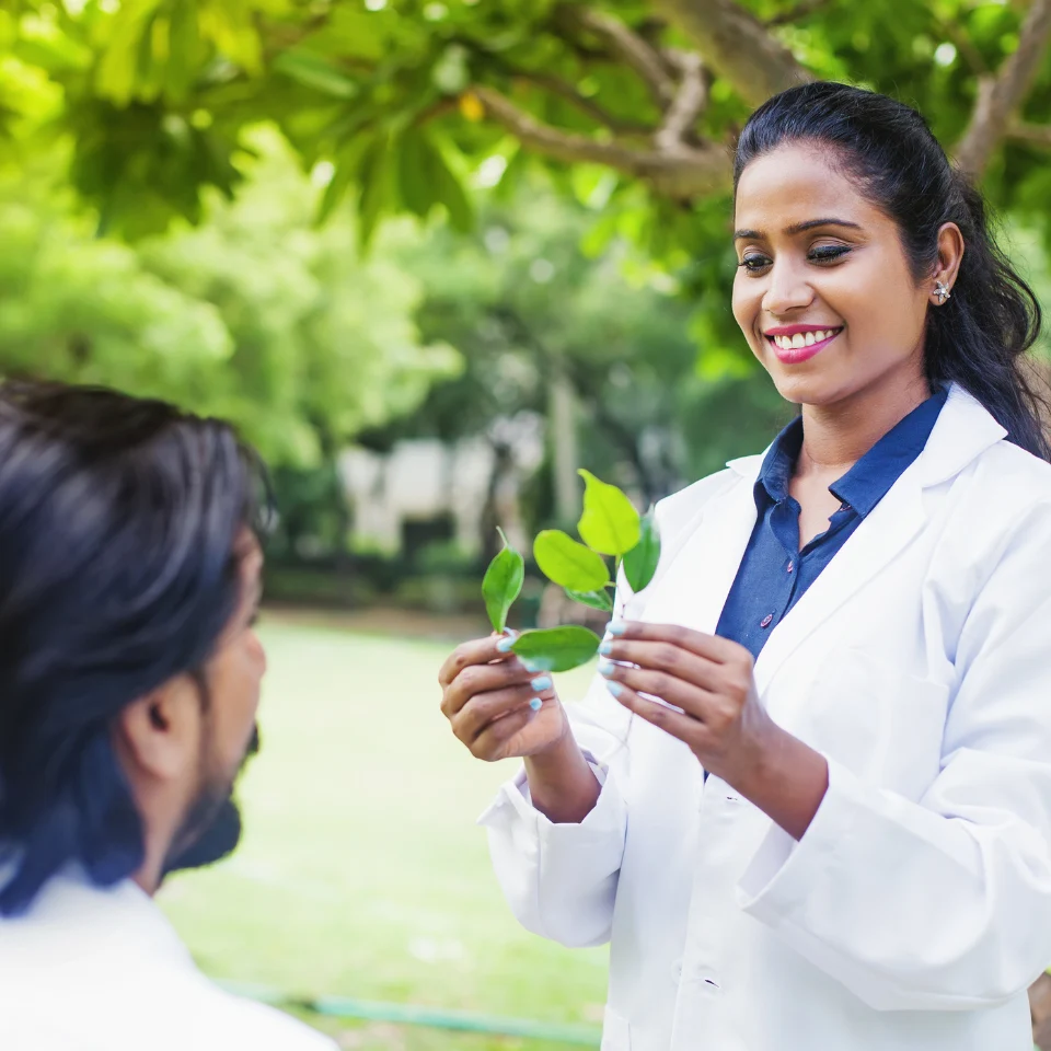 Smiling female scientist in a white lab coat holding green leaves and explaining them to a man in an outdoor garden setting, surrounded by lush greenery.