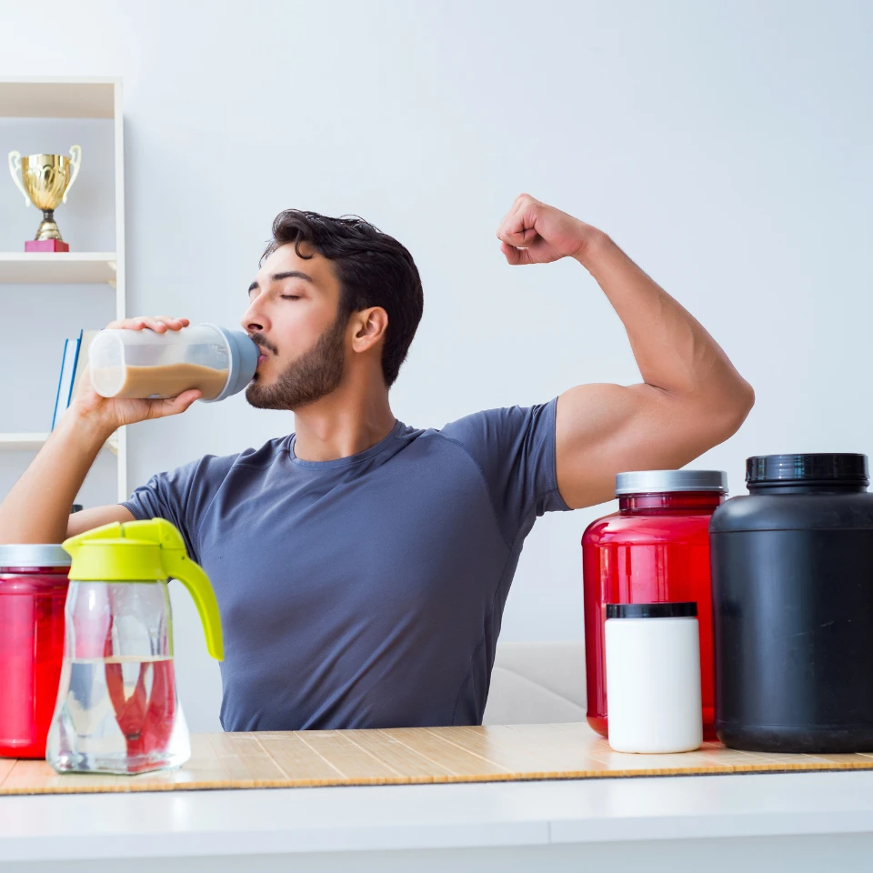 A fit young man flexing his arm while drinking a protein shake, sitting at a table with several large supplement containers and a water jug in front of him.