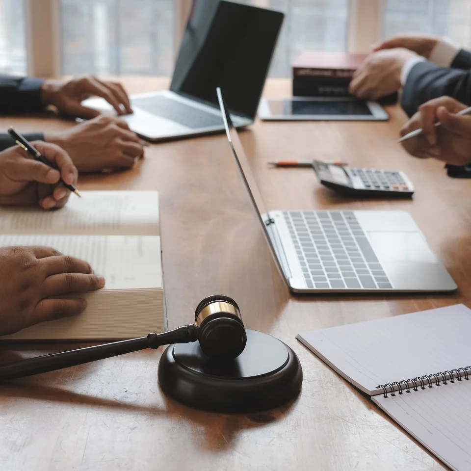 A group of people working at a table with open books, laptops, a calculator, and a judge’s gavel, suggesting a legal meeting or discussion.