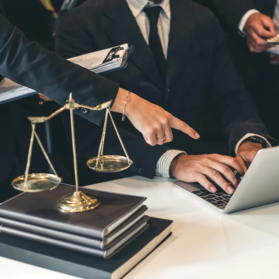 Legal professionals in formal attire discussing a case, with one person pointing at a laptop screen, a stack of law books, and justice scales on the desk.