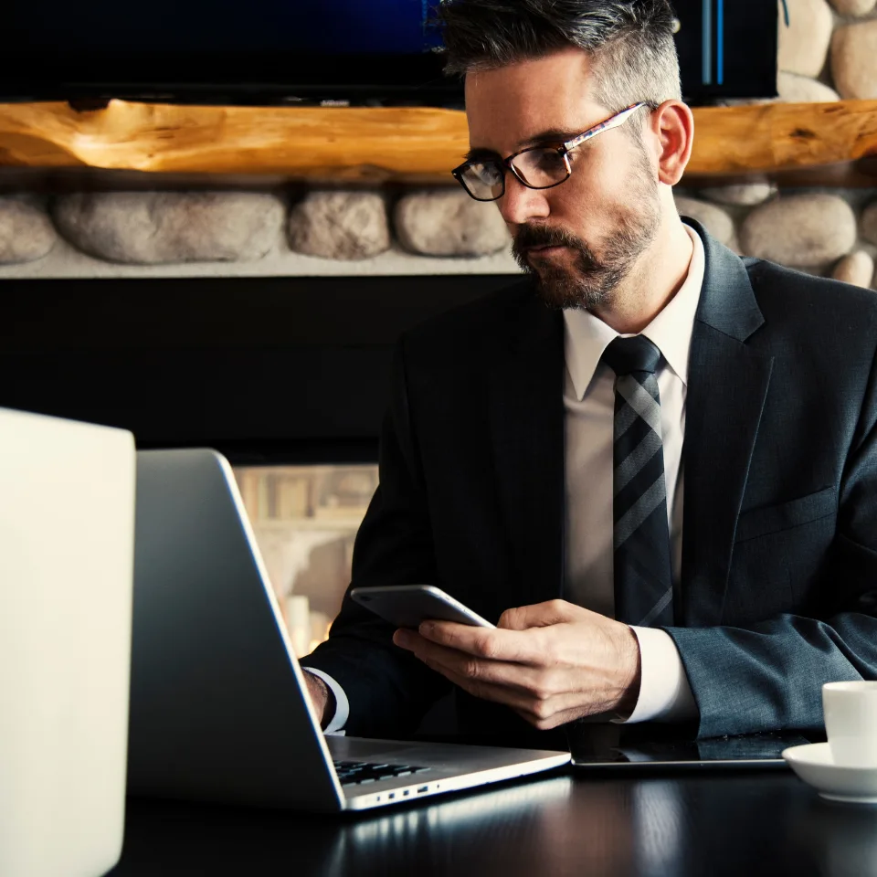 A serious-looking man in a suit and tie sitting at a desk, using a smartphone while working on a laptop, suggesting a legal professional or businessman at work.