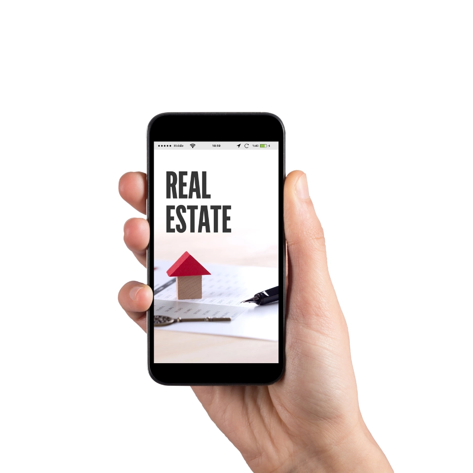 A hand holding a smartphone displaying a real estate app or website, with the words 'REAL ESTATE' and an image of a small house model with a red roof on the screen.