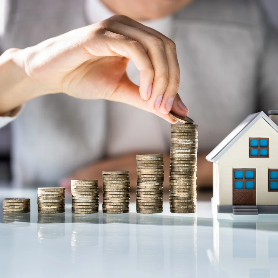  A person stacking coins in ascending order next to a small model house, symbolizing saving money or investing in real estate or property.