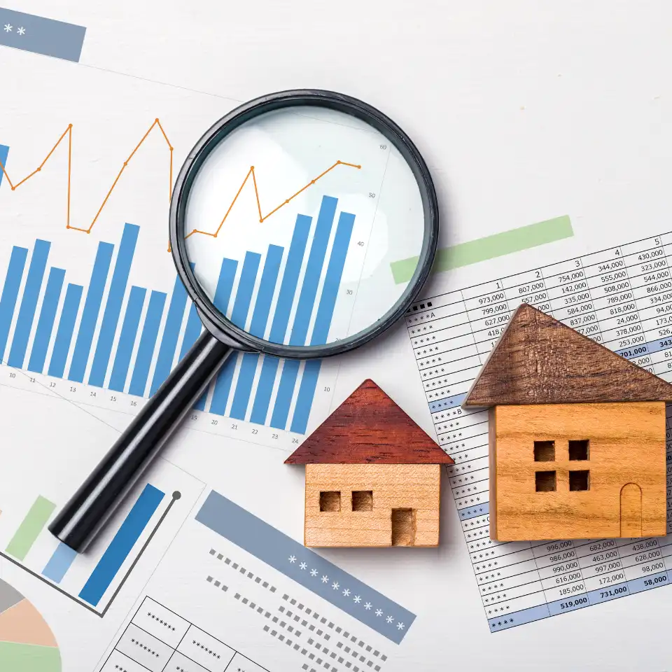 A magnifying glass placed over financial charts and graphs, next to two small wooden house models, representing real estate market analysis and investment research.