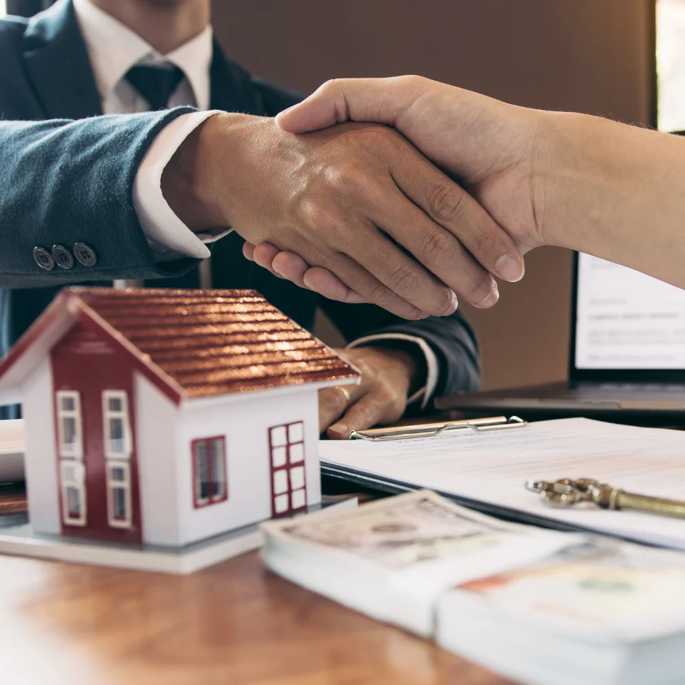 Two people shaking hands over a desk with real estate documents, money, and a model house, symbolizing a successful property deal or agreement.