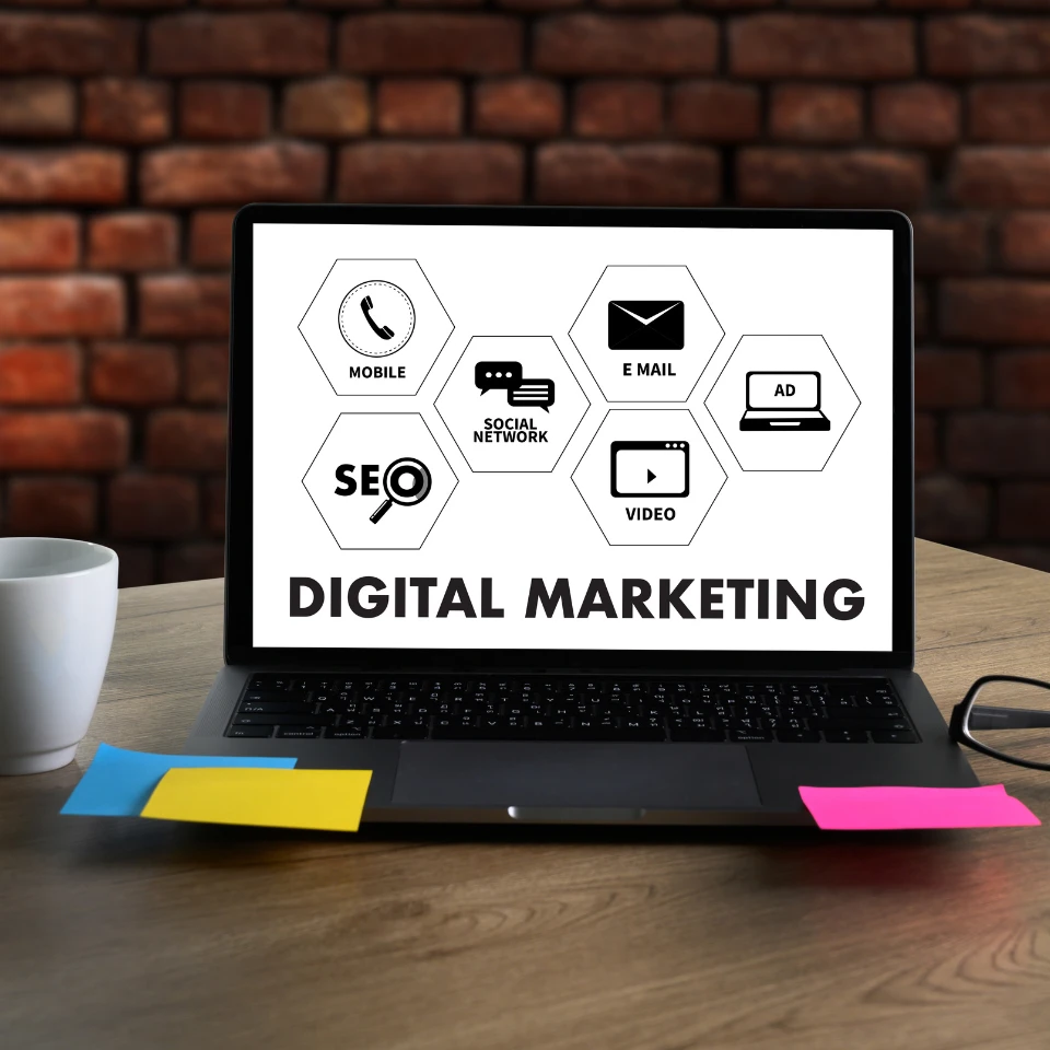 Laptop displaying DIGITAL MARKETING with hexagonal icons for SEO, mobile, email, social network, video, and ads on desk.