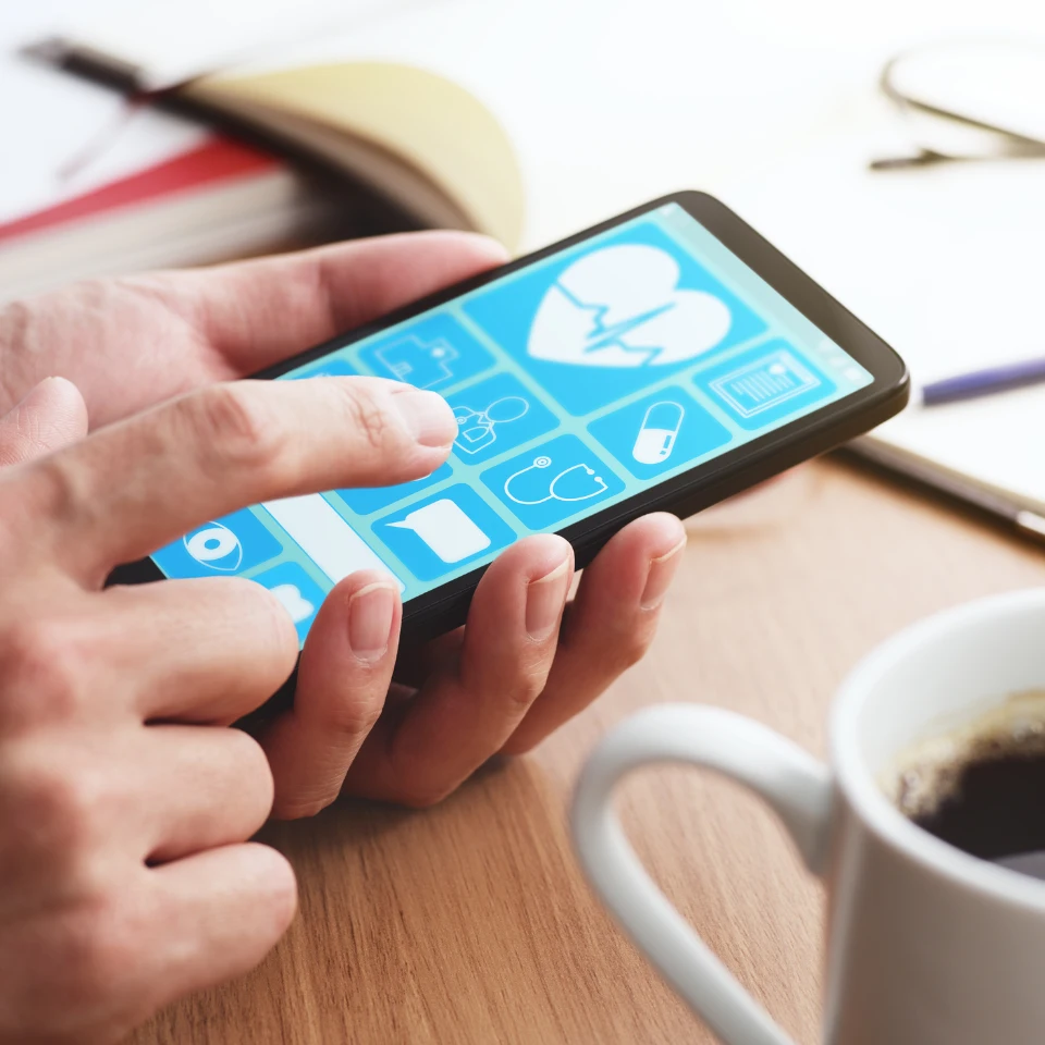 Hands holding tablet displaying medical app interface with health icons on wooden desk with coffee cup and notebook.