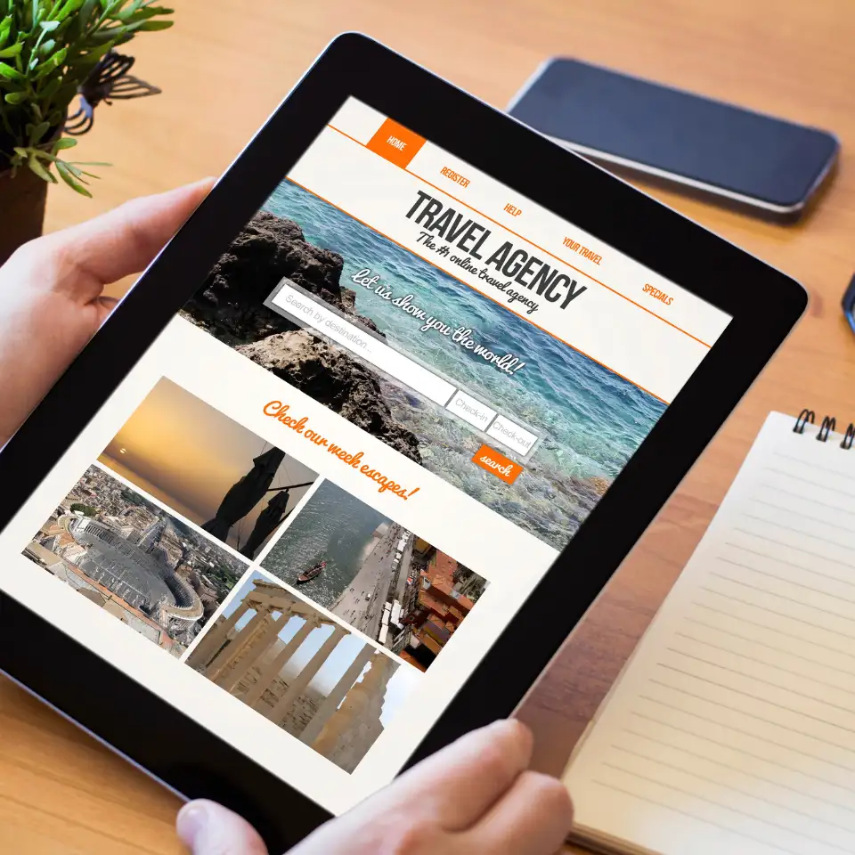 Person holding a tablet displaying a travel agency website with destination search options, scenic travel images, and promotional deals, placed on a wooden desk with a notebook and phone.