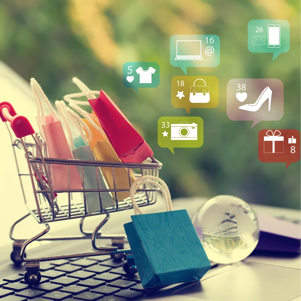 Mini shopping cart filled with colorful paper bags placed on a laptop keyboard, with floating digital icons representing online shopping, fashion, electronics, and messages.