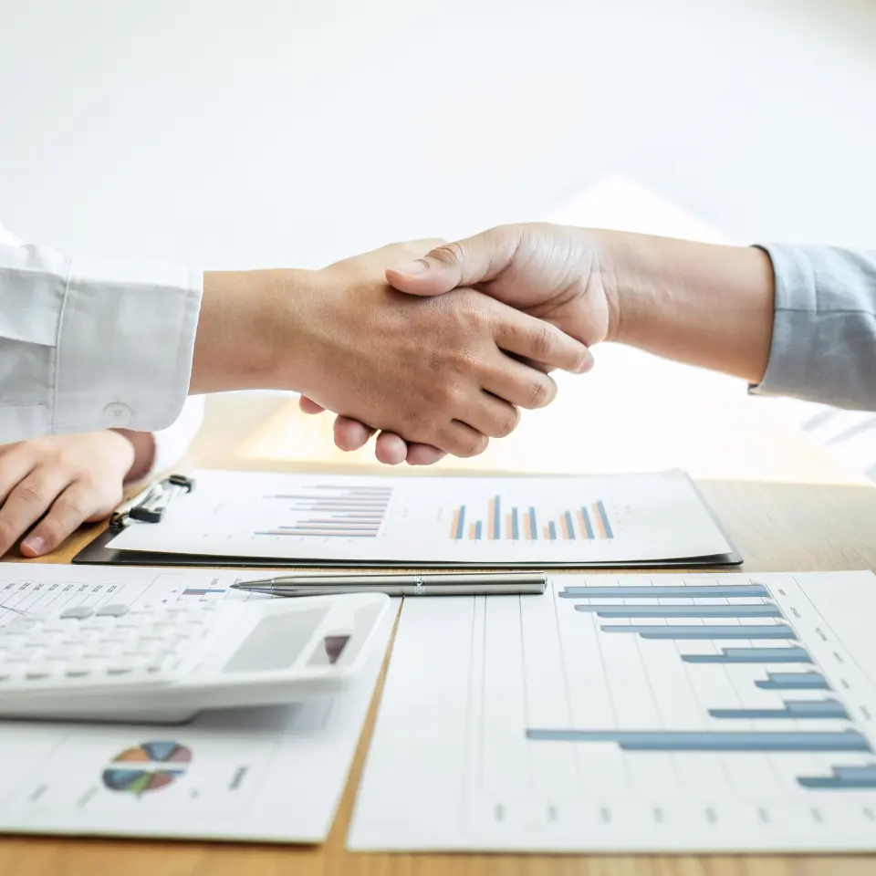 Two-business-professionals -shaking -hands-over-a-desk-with-charts-graphs-calculator-and-clipboard-symbolizing-a-successful-agreement-or-deal