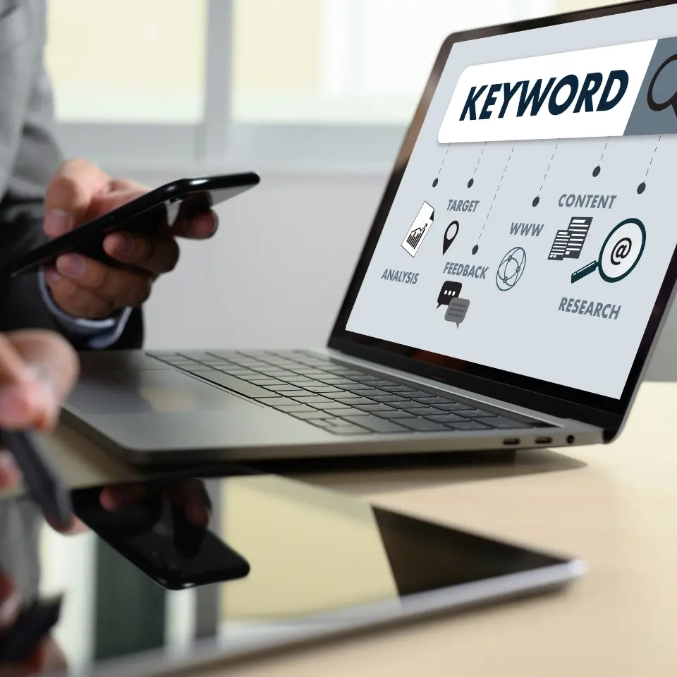 Person using a smartphone near a laptop screen displaying keyword research concepts like analysis, content, feedback, and targeting.