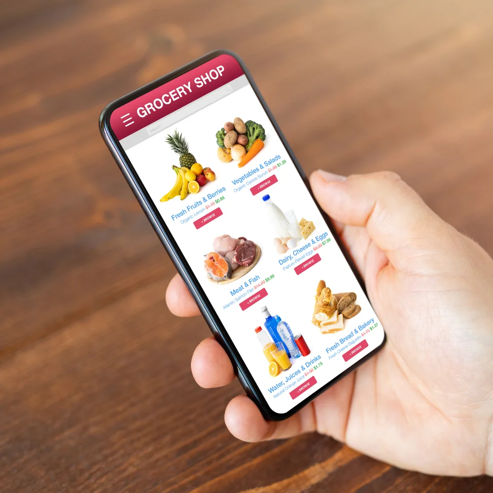 Hand holding a smartphone with an online grocery shopping app displaying categories like fruits, vegetables, dairy, and bakery.
