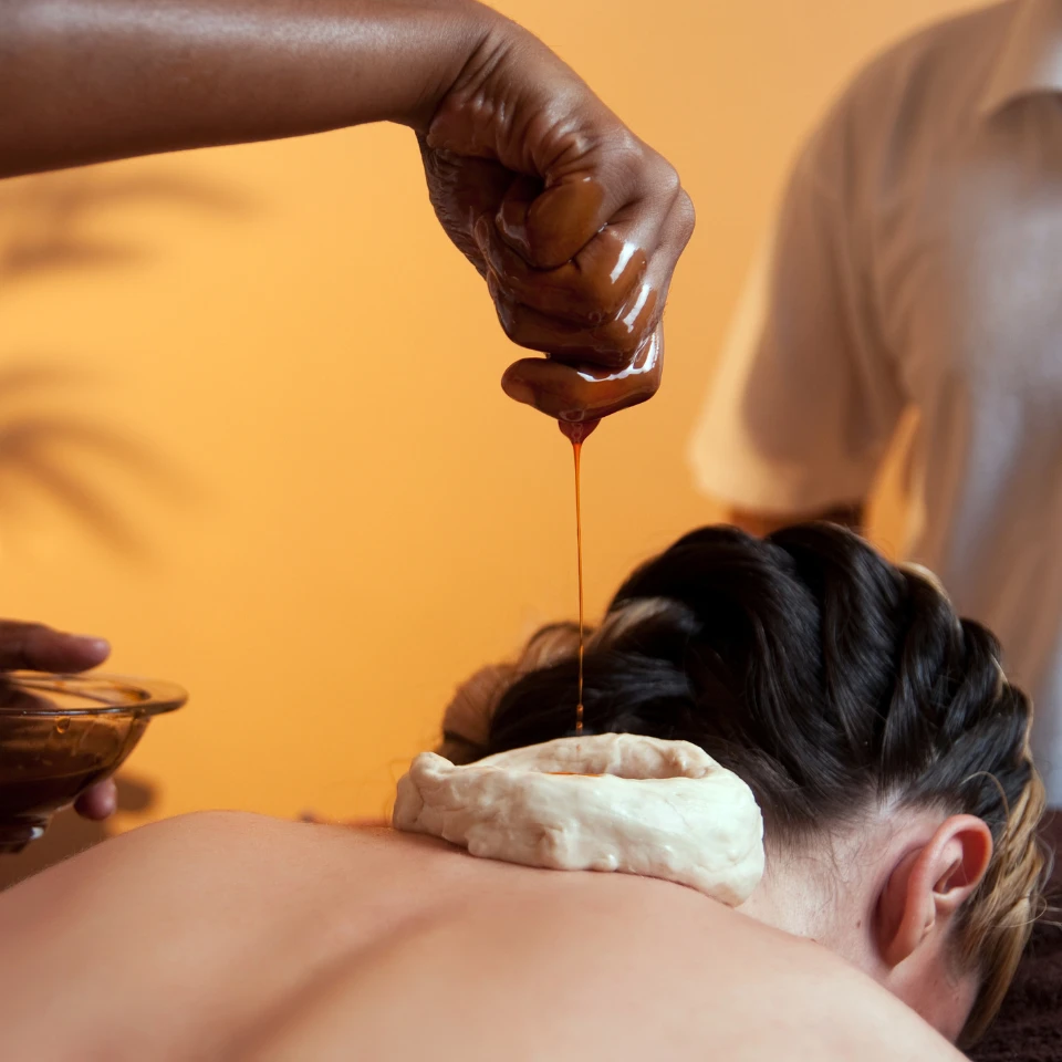 Close-up of an Ayurvedic therapy session where warm herbal oil is being poured into a dough dam placed on a woman's upper back for pain relief and relaxation.