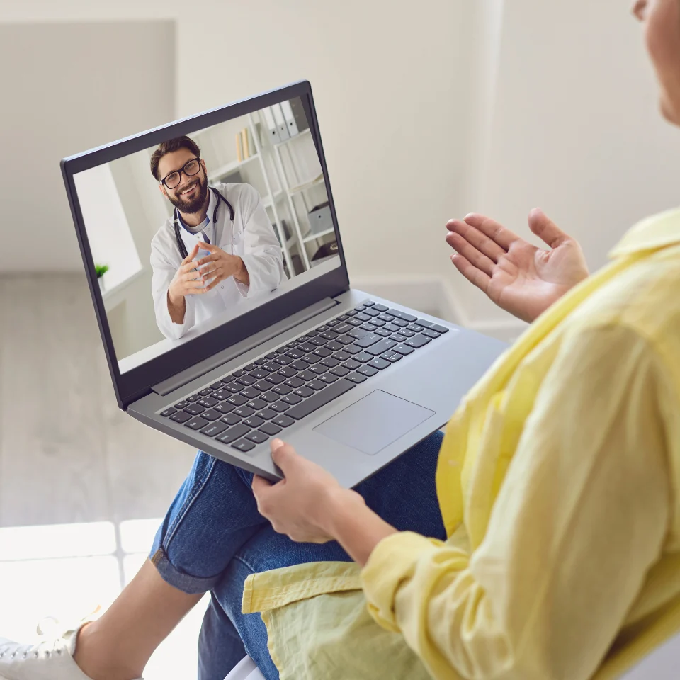 Woman sitting on a chair having an online video consultation with a smiling male doctor displayed on her laptop screen in a well-lit room.