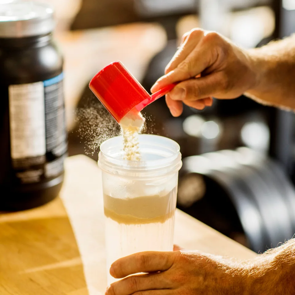 A person scooping protein powder into a clear shaker bottle with water, using a red scoop, at a wooden table in a gym or fitness setting.
