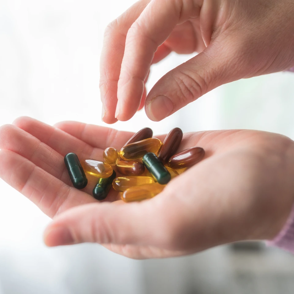 A person holding various colorful capsules and softgel supplements in one hand while picking one up with the other hand