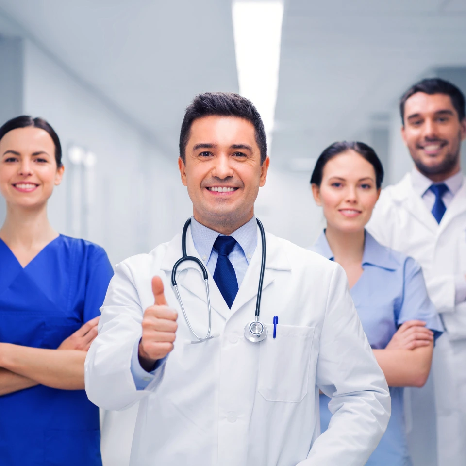 Smiling doctor in white coat giving thumbs up with medical team of four healthcare professionals in hospital corridor.