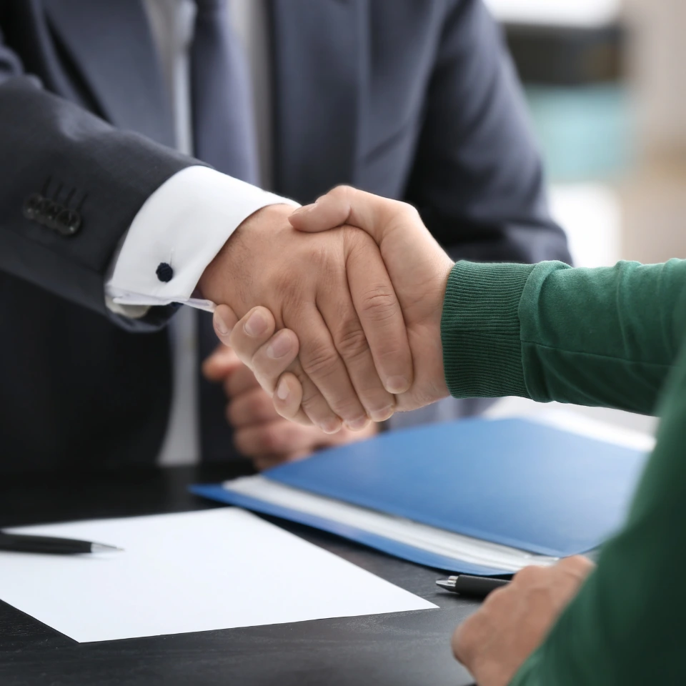 Close-up of a handshake between two people, one in a business suit and the other in casual clothing, with documents and a pen on the table, symbolizing a legal agreement or business deal.