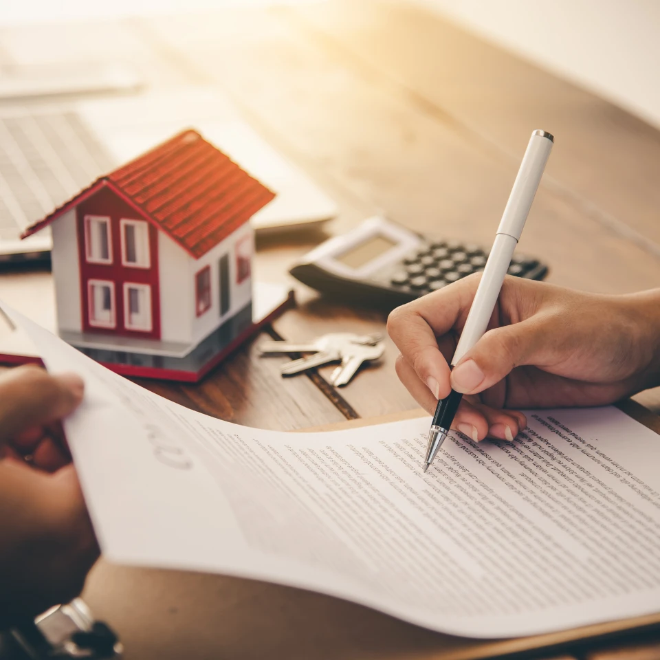 A person signing a real estate contract on a wooden desk with a model house, calculator, and keys in the background, symbolizing a property purchase or agreement.