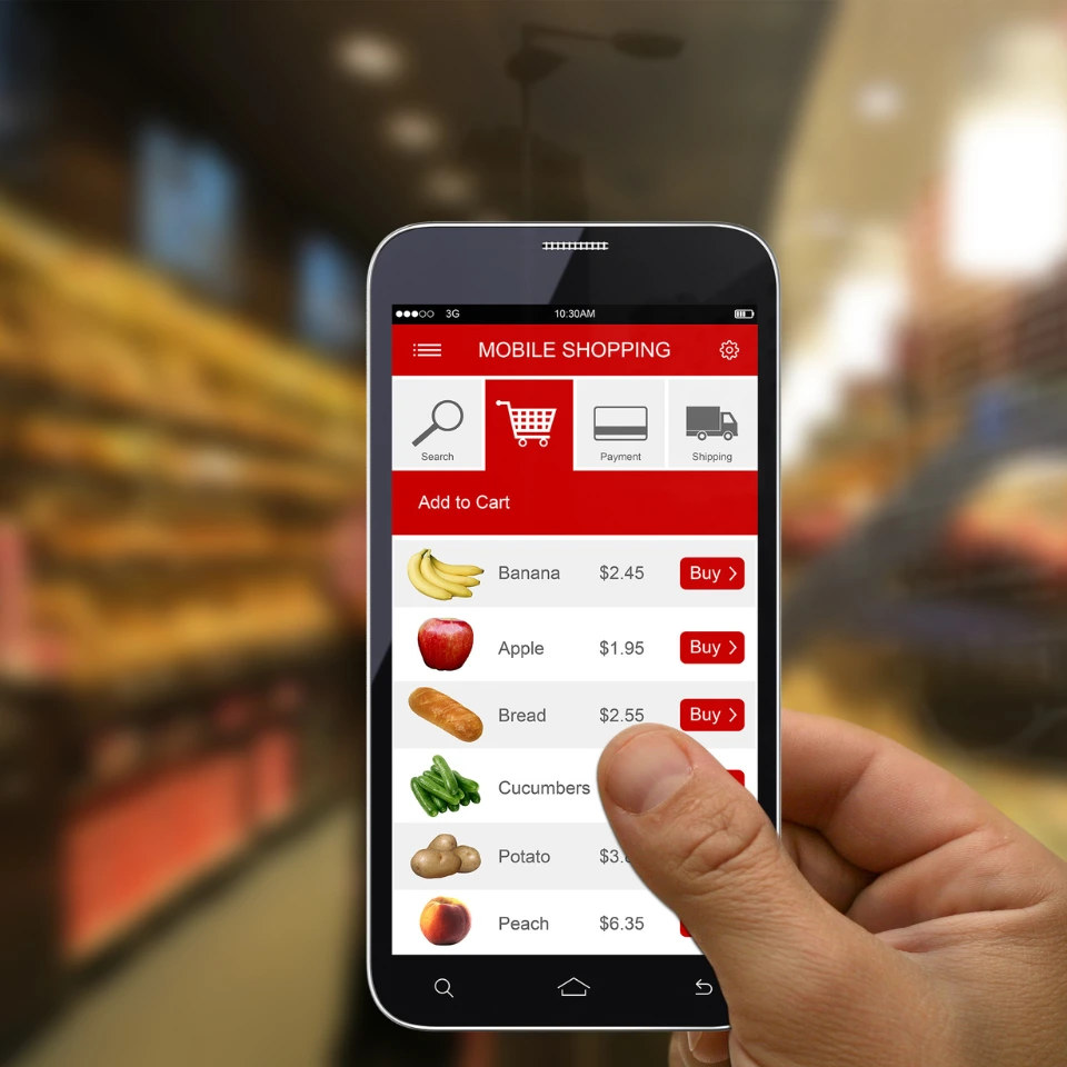 Hand holding smartphone with mobile shopping app displaying grocery items like banana, apple, bread with prices and buy buttons.