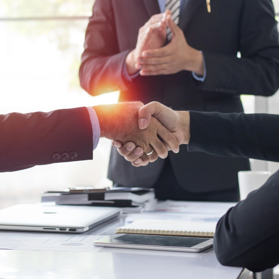 Business professionals shaking hands across desk with laptop and documents, sealing deal in bright office setting.