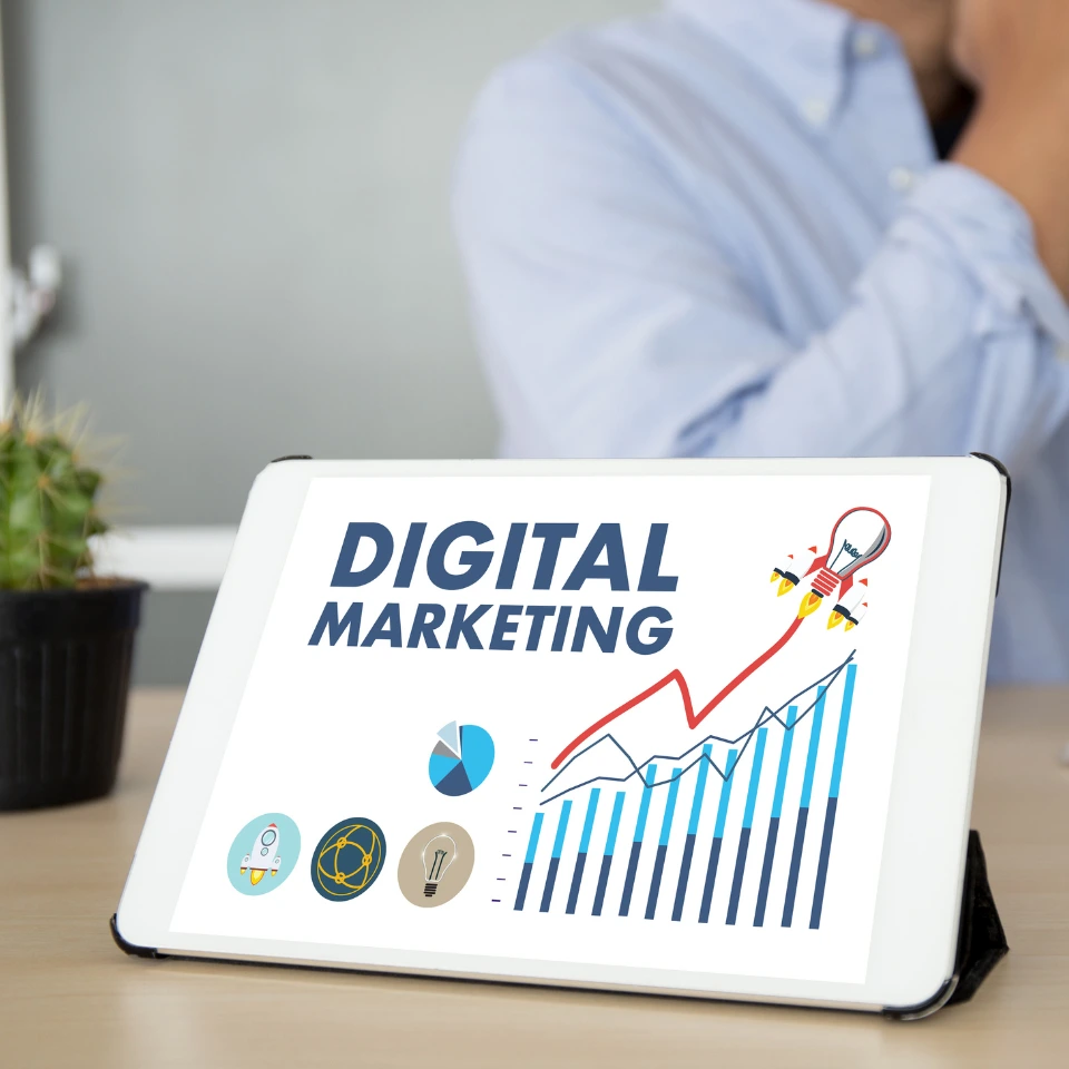 Tablet displaying DIGITAL MARKETING with growth chart, pie chart, and marketing icons on desk with blurred person behind.