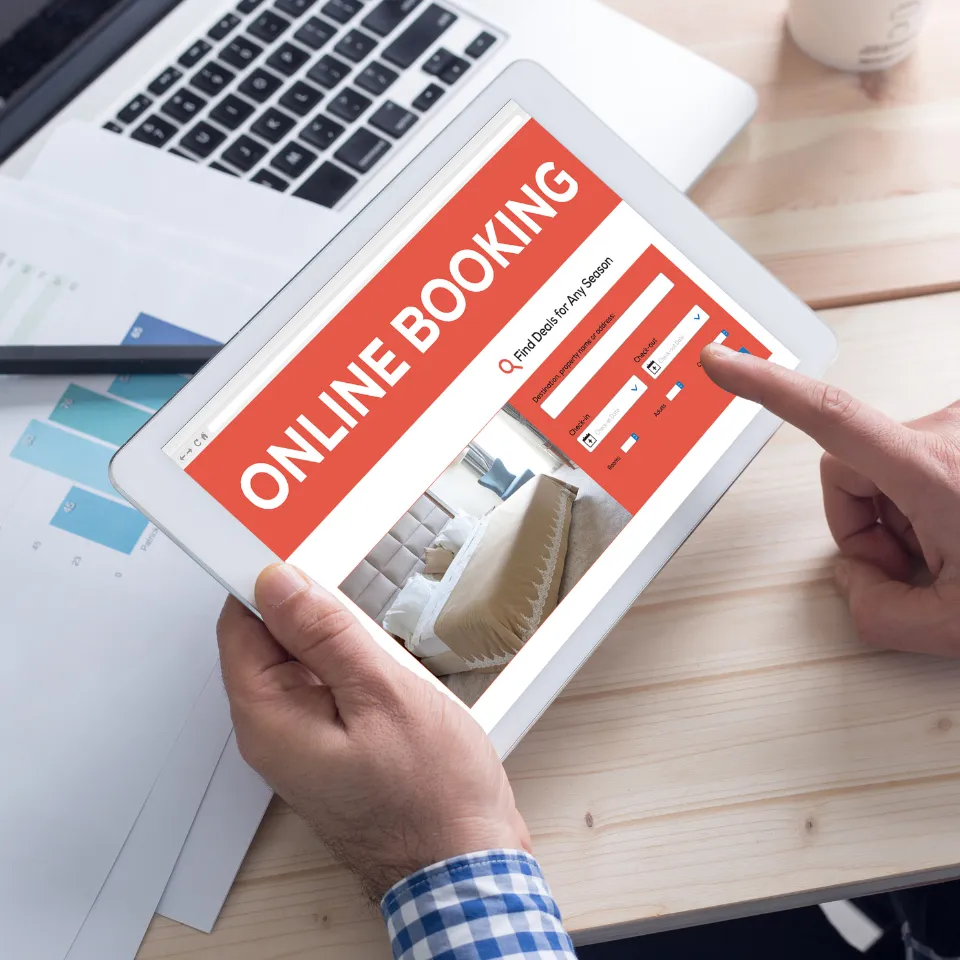 Person using a tablet to make an online hotel booking, with a reservation form and room image displayed on the screen, placed on a wooden desk with documents and a laptop.