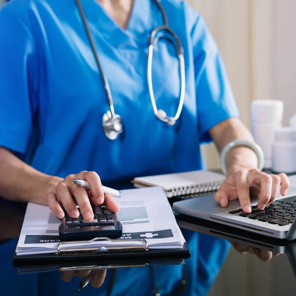 Medical professional in blue scrubs with stethoscope using calculator and laptop while reviewing charts and documents at desk.